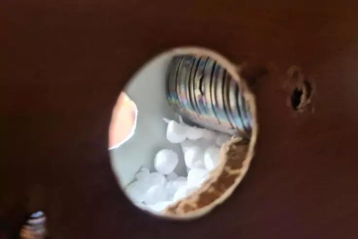 Rundes Loch in brauner Fläche, Blick in einen Hohlraum mit weißen Verpackungschips und einem silbernen, gewellten Rohr