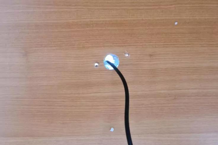 Helle Holzoberfläche mit rundem Loch mit silbernem Rand, schwarzem Kabel und drei kleinen silbernen Schrauben