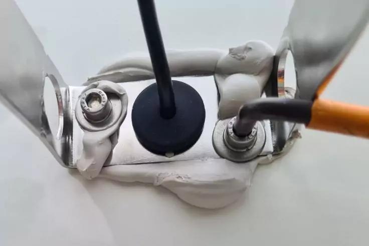 Nahaufnahme von zwei Metallklemmen mit angeschlossenen Kabeln (schwarz und orange), einer runden schwarzen Kabeldurchführung, weißer Dichtmasse und silbernen Halteblechen auf weißem Untergrund