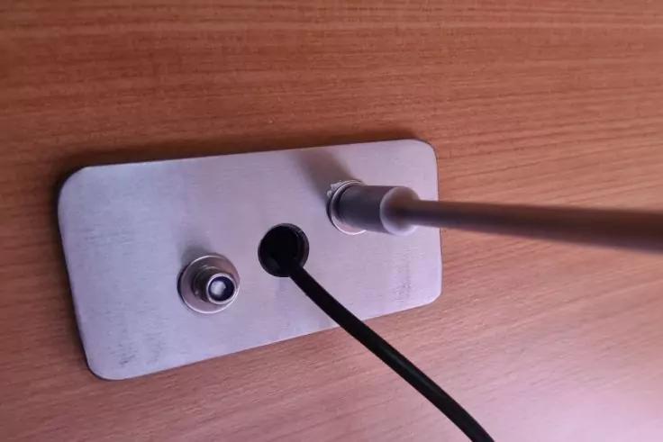 Rechteckige Metallplatte auf Holzoberfläche, zwei runde Anschlussbuchsen, ein schwarzes Kabel und eine silberne Stange