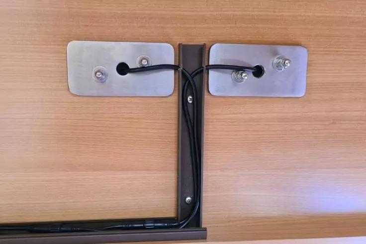 Holzoberfläche mit zwei rechteckigen Metallplatten, sichtbare Schrauben, schwarze Kabel durch Gummitüllen geführt, schwarzer Metallträger in der Bildmitte, schwarze Leitung entlang einer unteren Schiene