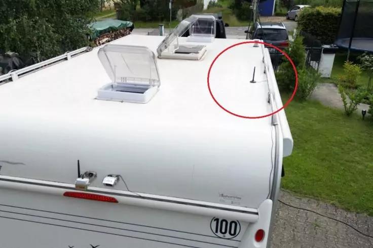 Dach eines weißen Wohnmobils, zwei geöffnete Dachluken mit transparenten Abdeckungen, schlanke Antenne nahe der rechten Kante, roter Kreis markiert die Antenne, Heck mit Rücklicht und Aufkleber "100", Rasen und Zufahrt im Hintergrund