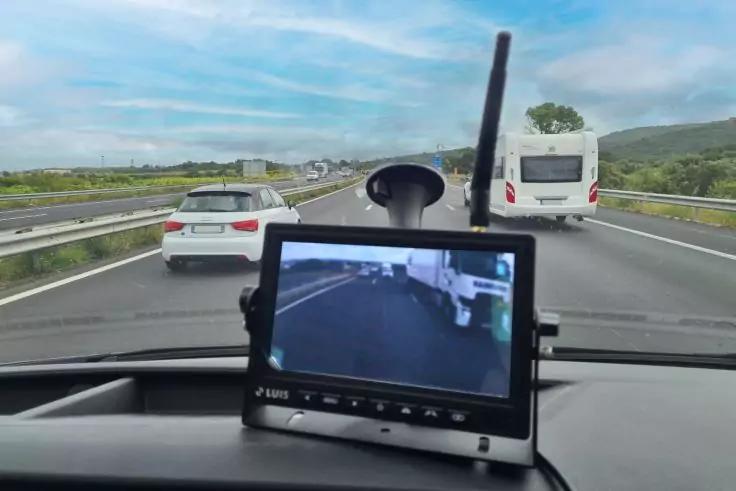Blick aus einem Fahrzeug auf eine Autobahn; im Vordergrund ein am Armaturenbrett montierter Monitor mit Kamerabild, davor ein weißer Van, links ein weißes Auto, Schutzplanken und hügelige Landschaft unter bewölktem Himmel