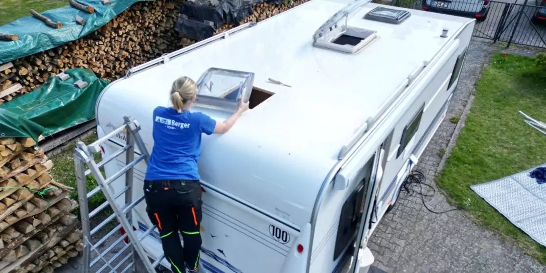 Person auf Leiter neben offenem Dachfenster eines wei&szlig;en Wohnwagens; daneben Stapel Brennholz, Rasenfl&auml;che und Einfahrt