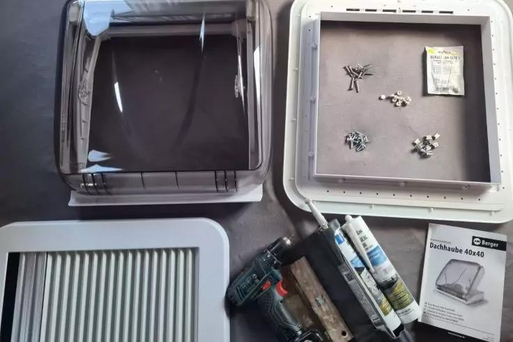 Zwei rechteckige Kunststoffrahmen (schwarz, wei&szlig;), wei&szlig;e Schale mit Schrauben und kleinen Metallteilen, gr&uuml;ner Akkuschrauber, zwei Kartuschen, Bedienungsanleitung auf grauem Untergrund