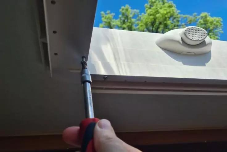 Hand mit Schraubendreher an einer ge&ouml;ffneten Dachluke; sichtbares Metallgest&auml;nge, L&uuml;ftungsgitter und blauer Himmel mit B&auml;umen