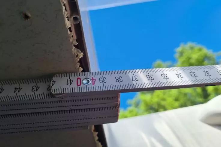 Ausgezogenes Metallma&szlig;band mit Zentimeter-Skala vor einer Dachkante, blauer Himmel und gr&uuml;ner Baum im Hintergrund