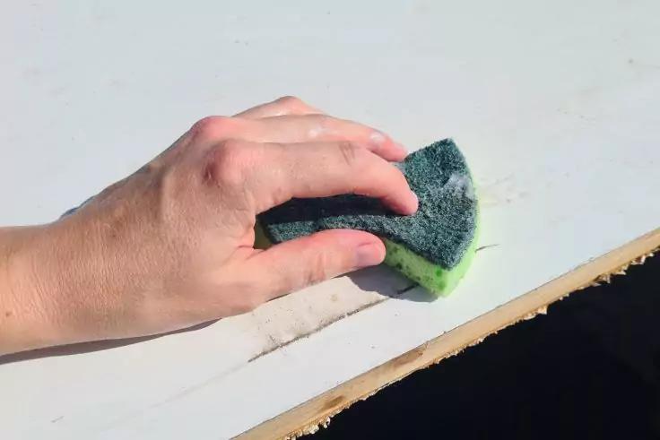 Hand h&auml;lt gr&uuml;nen Schwamm auf wei&szlig;er Oberfl&auml;che mit sichtbarem Schmutz