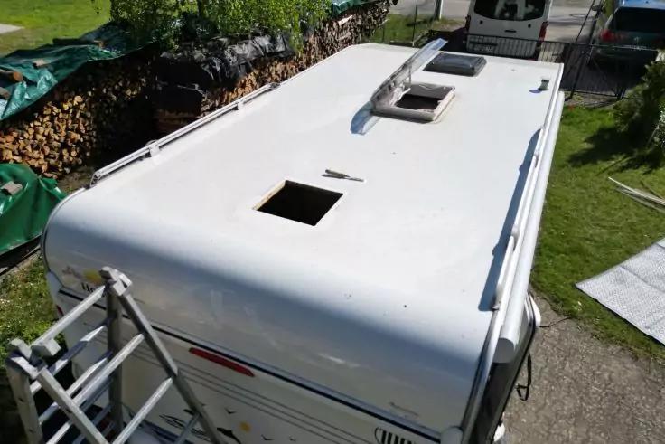 Dach eines wei&szlig;en Wohnwagens mit zwei rechteckigen &Ouml;ffnungen, Leiter an der Seite, Holzstapel, Rasen und wei&szlig;es Fahrzeug im Hintergrund