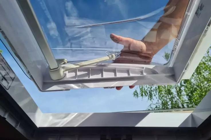 Ge&ouml;ffnetes Dachfenster von innen, Hand h&auml;lt den Griff, blauer Himmel und Baumzweige drau&szlig;en