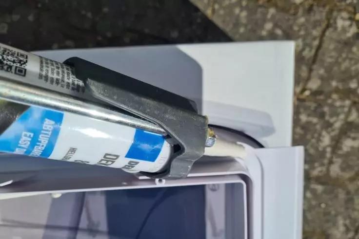 Kartuschenpistole mit eingelegter Dichtstoffkartusche (blaues Etikett) auf wei&szlig;er Verpackung neben einem Smartphone in seinem Fach, im Hintergrund Pflaster