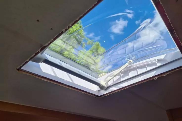 Rechteckiges Dachfenster mit wei&szlig;em Rahmen; durch die Glasscheibe sichtbarer blauer Himmel mit Wolken und Kondensspur sowie gr&uuml;ne Baumzweige; Innendecke mit sichtbarem Putzrand
