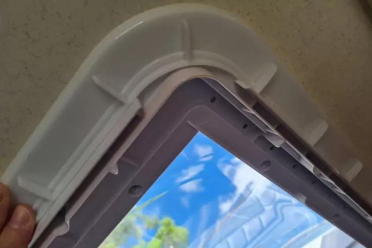 Wei&szlig;er Kunststoffrahmen einer ge&ouml;ffneten rechteckigen &Ouml;ffnung, blauer Himmel mit Wolken und Baumbl&auml;ttern sichtbar, Hand h&auml;lt den Rahmen, beige Innenwand