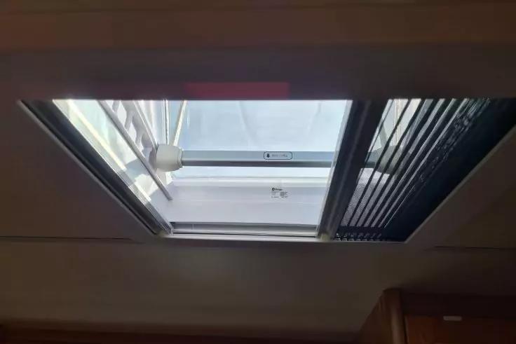 Rechteckiges Dachfenster mit Glas und Rahmen, rechts teilweise geschlossene Jalousie, blauer Himmel mit Wolken drau&szlig;en, Deckenrand und Holzverkleidung unten sichtbar