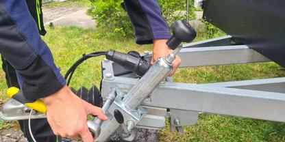 Zwei H&auml;nde an der Metallkupplung eines Bootsanh&auml;ngers; Anh&auml;ngerrahmen und Bootsrumpf sichtbar, Gras im Hintergrund
