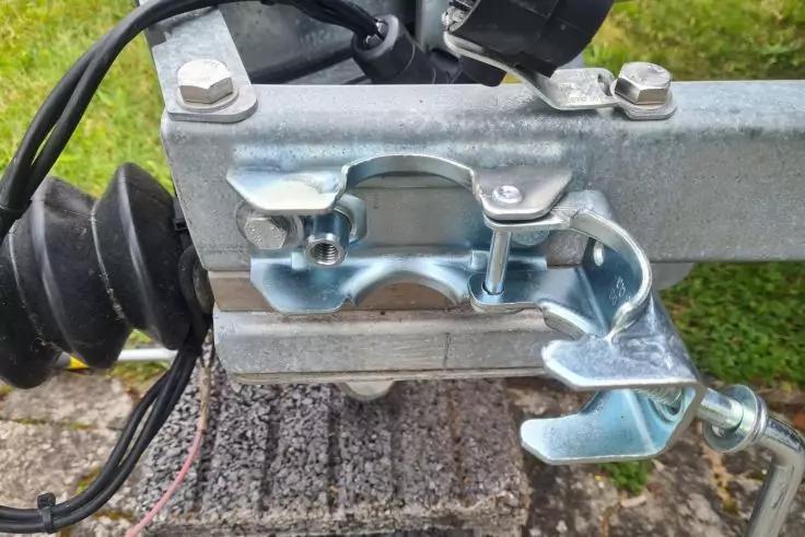 Nahaufnahme einer metallischen Anh&auml;ngerkupplung mit Hebel und Klammer, Gummimanschette, Kabeln, Metallrahmen, Betonplatte und Gras im Hintergrund