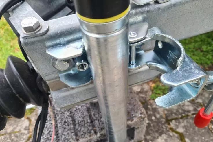 Silbernes senkrechtes Metallrohr, daran befestigte verzinkte Metallhalterung mit Hebel und Verriegelungsb&uuml;gel, links schwarzes Kunststoffrad, rechts roter Kunststoffgriff an d&uuml;nnem Kabel, Pflastersteine und Gras im Hintergrund