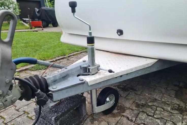 Vorderer Bereich eines wei&szlig;en Anh&auml;ngers auf Pflaster: verzinkte Deichsel mit montiertem St&uuml;tzrad und Kurbel, angeschlossenes Kabel, Gras und roter Gegenstand im Hintergrund