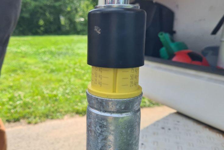 Nahaufnahme eines verzinkten Metallrohrs mit gelber Kunststoffh&uuml;lse mit Skalenmarkierungen und schwarzer Kappe; links Rasen, rechts offene Fahrzeugheckklappe mit sichtbaren Werkzeugen