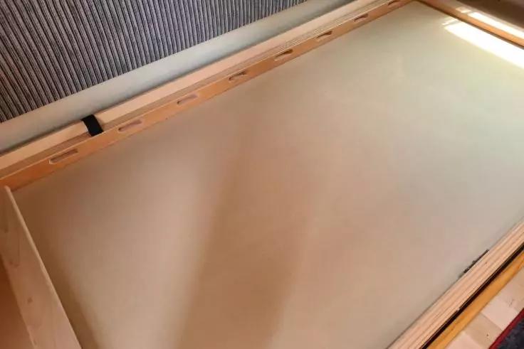 Holz-Bettenrahmen mit beiger Matratzenunterlage, grauer gepolsterter Kopfteilkante und Lichteinfall oben rechts