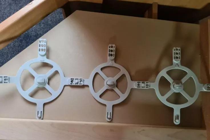 Drei runde wei&szlig;e Scheiben mit radialen Speichen und kurzen Laschen nebeneinander auf einer braunen Holzfl&auml;che, dar&uuml;ber ein Holzrahmen sichtbar