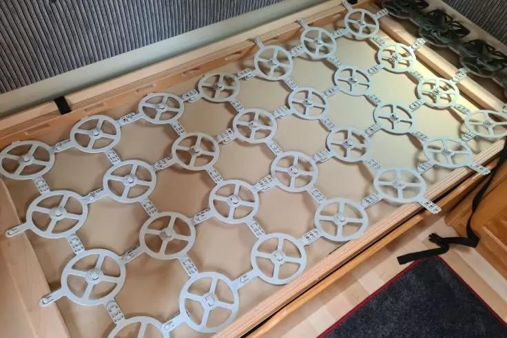 Holzrahmen mit miteinander verbundenen grauen Kunststoffringen in wiederholendem Gittermuster, schwarze B&auml;nder, Teppichkante und Heizk&ouml;rper im Hintergrund