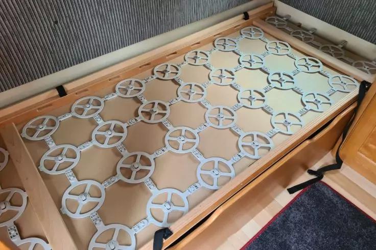 Bettgestell aus Holz ohne Matratze mit Gitter aus beigen/wei&szlig;en runden Plastikplatten; Holzschrank rechts; gestreifte Wand im Hintergrund; dunkler Teppichboden und schwarzer Riemen am Rahmen