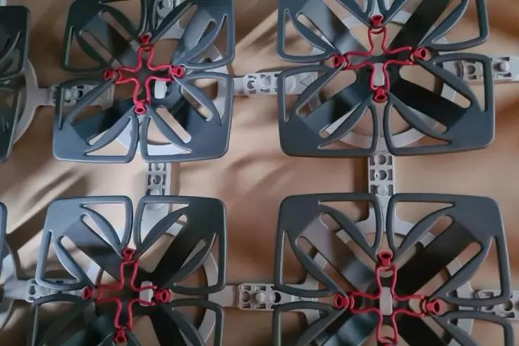 Mehrere dunkelgraue quadratische Kunststoffrahmen, jeweils mit vier gebogenen Bl&auml;ttern und roten Mittelteilen, auf beigem Stoff angeordnet