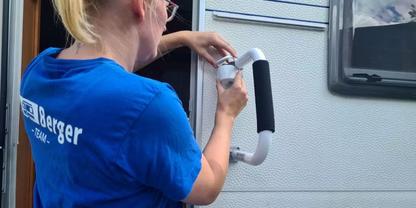 Person mit blond zusammengebundenen Haaren und blauem T-Shirt h&auml;lt einen wei&szlig;en Haltegriff an der Au&szlig;enseite eines Wohnmobils neben einer T&uuml;r