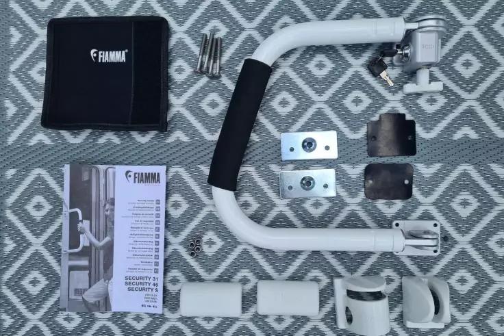 Auf einem grauen Untergrund mit wei&szlig;em Rautenmuster liegen mehrere Teile: ein wei&szlig;er U-f&ouml;rmiger Metallb&uuml;gel mit schwarzer Schaumstoffummantelung, wei&szlig;e Kunststoffh&uuml;lsen und -schellen, zwei rechteckige Metallplatten, zwei schwarze Gummipads, eine schw