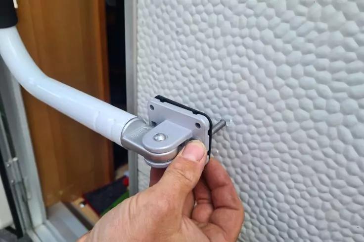 Hand h&auml;lt eine kleine rechteckige Metallplatte mit schwarzem Rand an ein wei&szlig;es, strukturiertes T&uuml;rpaneel neben einem gebogenen wei&szlig;en Metallarm