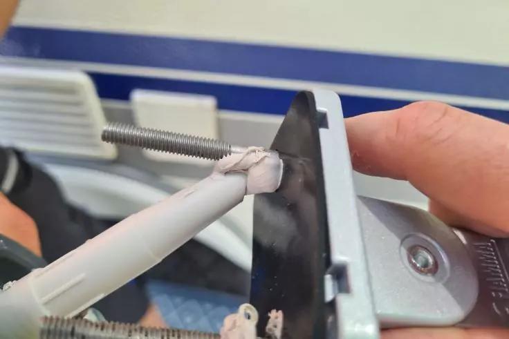 Rechte Hand h&auml;lt ein schwarzes Kunststoffteil mit einer Metall-Gewindestange und einem wei&szlig;en Kunststoffstab, an der Verbindungsstelle wei&szlig;e Dichtmasse; rechts eine silberne Schraube, unscharfer blau-wei&szlig;er Hintergrund