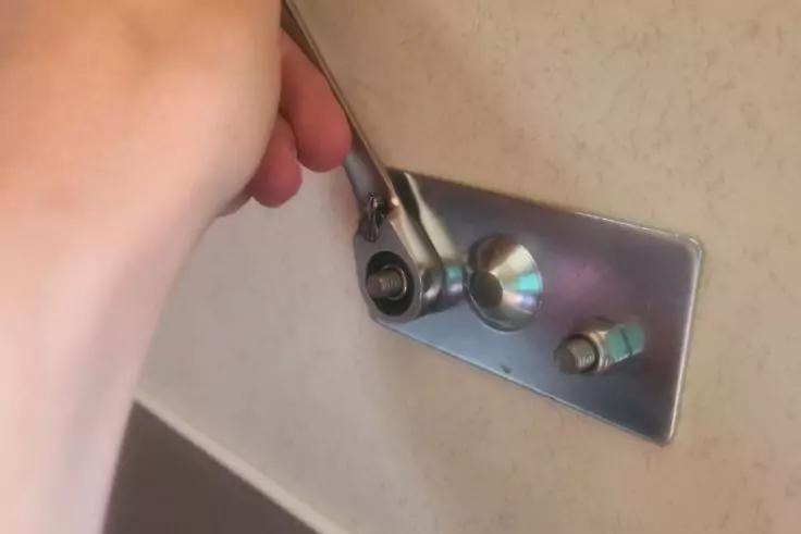 Hand h&auml;lt Ratschenschl&uuml;ssel an sechskantiger Schraube auf einer rechteckigen Metallplatte an einer Wand; zwei weitere Schrauben sichtbar