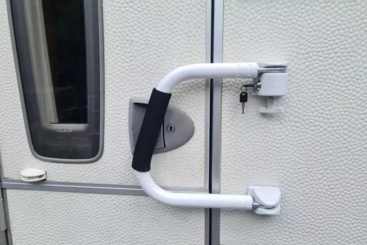 U-f&ouml;rmiger wei&szlig;er T&uuml;rgriff mit schwarzer Polsterung an einer hellen, strukturierten T&uuml;r; rechts ein Zylinderschloss mit Schl&uuml;ssel; links ein schmales Fenster