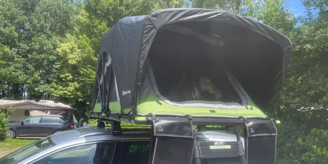 Auto mit gr&uuml;n-schwarzem aufgeklapptem Dachzelt auf dem Dach, Leiter h&auml;ngt bis zum Boden, im Hintergrund B&auml;ume und ein geparkter Wohnwagen