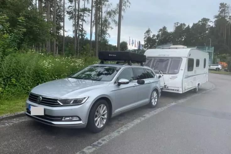 Silberner Kombi mit schwarzem Dachkoffer, an wei&szlig;en Wohnwagen angeh&auml;ngt, am Fahrbahnrand vor einem Wald geparkt