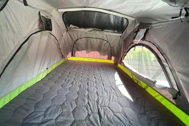 Innenansicht eines Zeltes mit grauer gesteppter Matratze, hellgr&uuml;ner Kante, Stoffw&auml;nden, seitlichen Fenster&ouml;ffnungen und ge&ouml;ffneten Rei&szlig;verschl&uuml;ssen