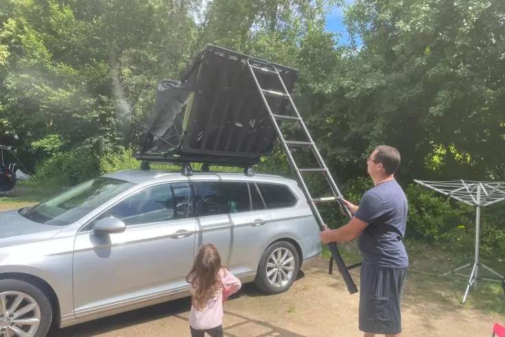 Silberner Kombi mit aufgeklapptem Dachzelt; eine erwachsene Person h&auml;lt eine Leiter, ein Kind steht daneben; B&auml;ume im Hintergrund