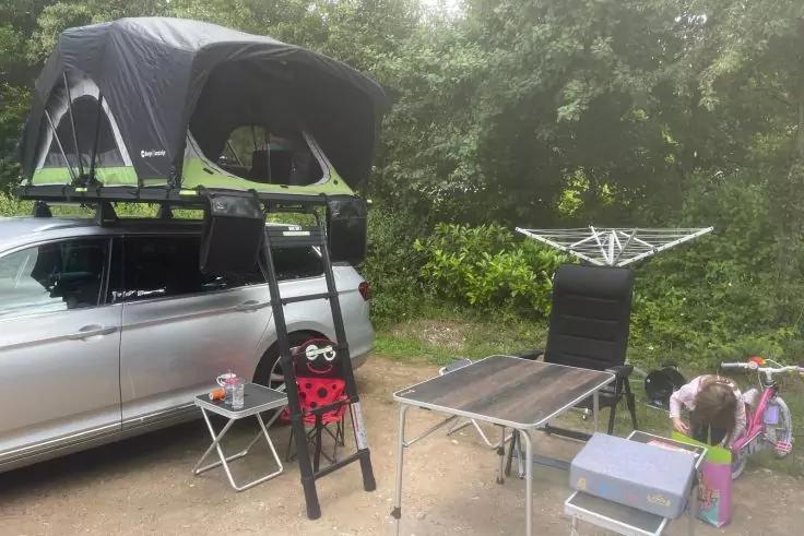 Silberner Kombi mit aufgeklapptem Dachzelt und Leiter, kleiner Klapptisch mit Getr&auml;nkedose, weiterer Tisch mit Stuhl, Kiste und Kinderwagen vor B&uuml;schen