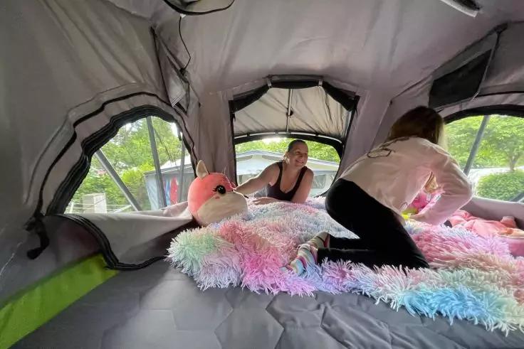 Zwei Personen in einem offenen Zelt auf einer bunten, flauschigen Decke mit einem gro&szlig;en Pl&uuml;sch-Einhorn; Zeltfenster zeigen B&auml;ume drau&szlig;en