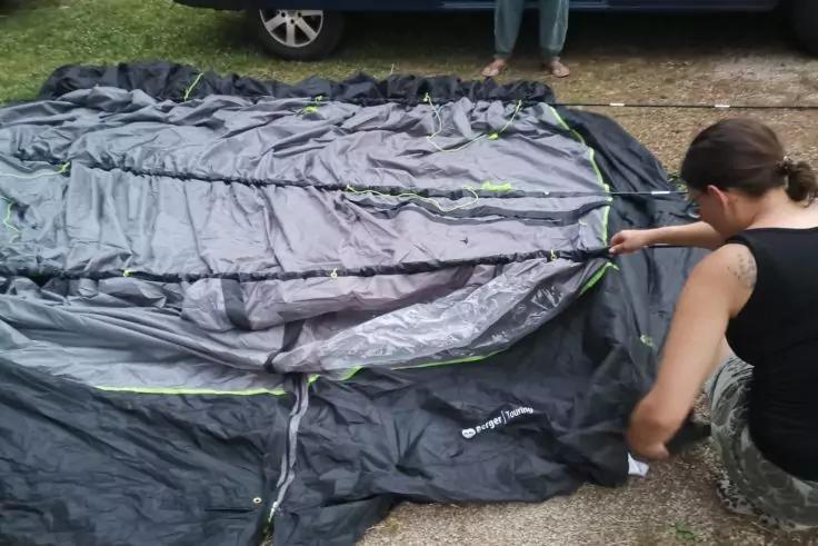 Person kniet rechts und fasst einen gro&szlig;en dunkelgrauen Stoff mit gr&uuml;nen Ziern&auml;hten und Rei&szlig;verschl&uuml;ssen an, der auf Kiesboden ausgebreitet liegt; blaues Auto mit sichtbaren Reifen im Hintergrund; Gras links