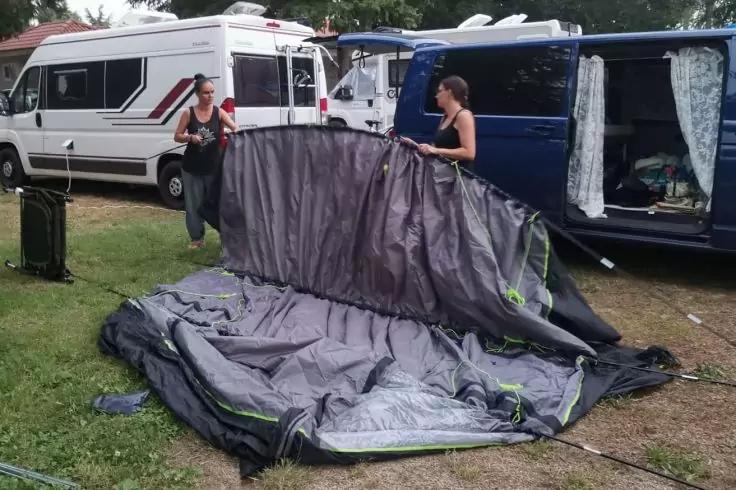 Zwei Frauen neben einem auf dem Boden ausgebreiteten grauen Zeltstoff zwischen einem wei&szlig;en Wohnmobil und einem blauen Van, Gras im Vordergrund