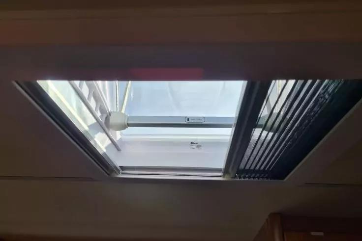 Rechteckiges Dachfenster in der Zimmerdecke, eine Glasscheibe gekippt, Metallrahmen, rechts Lamellenjalousie, links runde Deckenleuchte, heller Himmel sichtbar