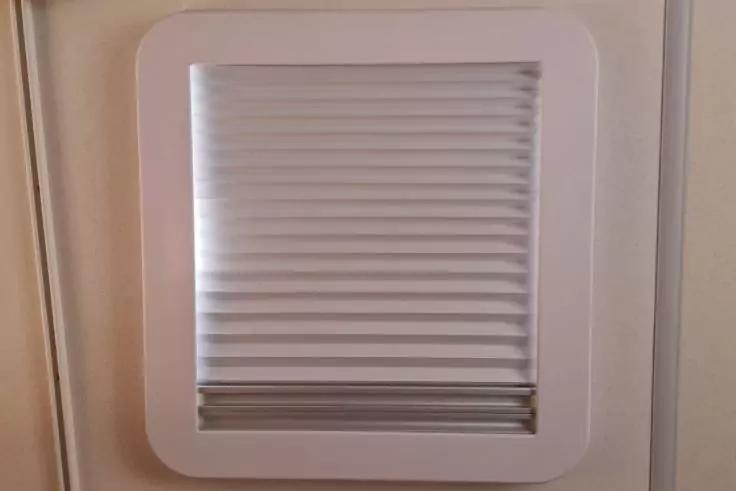 Quadratischer weißer Rahmen an beige Wand, darin ein hellgraues Plissee mit horizontalen Falten