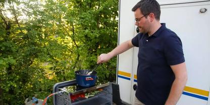 Mann mit Brille und dunklem Poloshirt r&uuml;hrt mit einem L&ouml;ffel in einem Topf auf einem tragbaren Camping-Gasherd neben einem wei&szlig;en Wohnmobil, B&auml;ume im Hintergrund