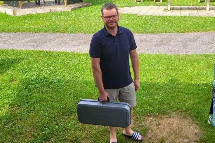 Mann steht auf Gras neben einem Gehweg, h&auml;lt einen grauen Hartschalenkoffer in der rechten Hand, tr&auml;gt dunkles Poloshirt, helle Shorts und schwarze Latschen mit wei&szlig;en Streifen, schaut in die Kamera und l&auml;chelt