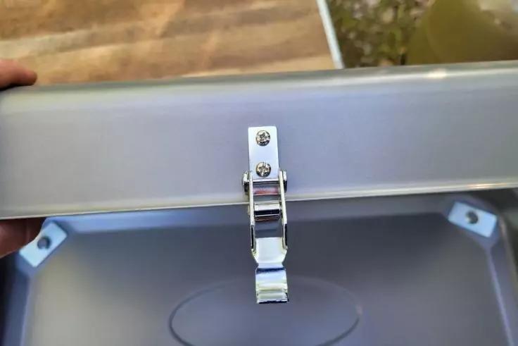 Ge&ouml;ffnete silberne Metallbox mit zentralem silbernem Verschlussriegel, Innenboden mit ovaler Vertiefung, zwei innere Eckbeschl&auml;ge, oben links von einer Hand gehalten, unscharfer Holztisch und gr&uuml;nes Objekt im Hintergrund