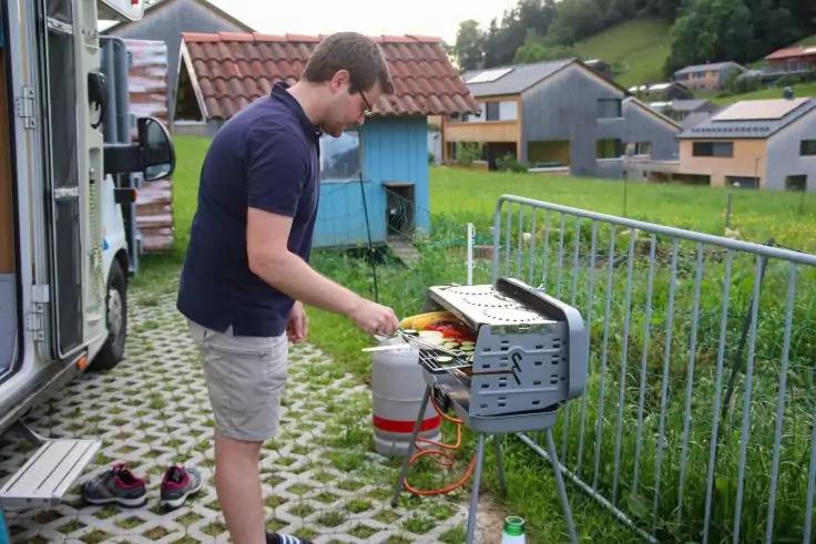 Eine Person steht an einem offenen tragbaren Gasgrill neben einer Propangasflasche; im Hintergrund Wohnmobil, Schuhe auf dem Boden, Gel&auml;nder, H&auml;user und Wiese
