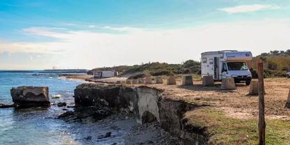 Wei&szlig;es Wohnmobil auf sandig-grasigem K&uuml;stenstreifen neben Felsvorsprung, Meer und Wolken am Himmel