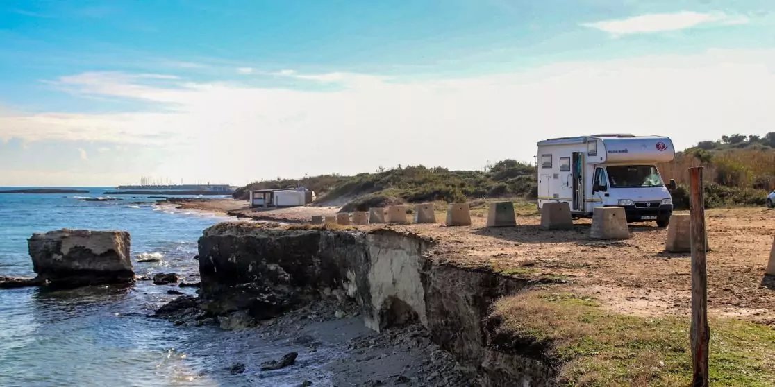 Wei&szlig;es Wohnmobil auf sandig-grasigem K&uuml;stenstreifen neben Felsvorsprung, Meer und Wolken am Himmel